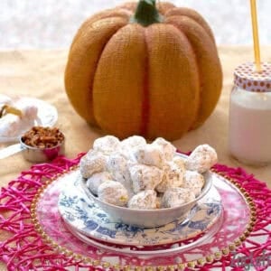 A bowl of Pumpkin and Pecan Polvorones coated in powdered sugar plated next to a measuring cup of pecans, a large decorative pumpkin, and a glass of milk with a straw
