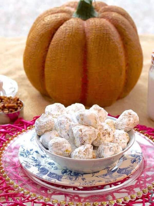 A bowl of Pumpkin and Pecan Polvorones coated in powdered sugar plated next to a measuring cup of pecans, a large decorative pumpkin, and a glass of milk with a straw