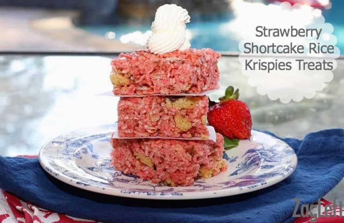 Three Strawberry Shortcake Rice Krispies Treats stacked on a plate topped with whipped cream with a strawberry on the side of the plate