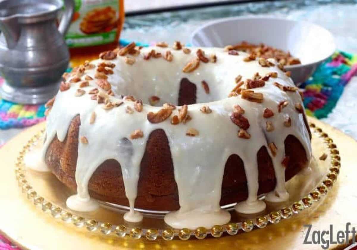 Pumpkin Bundt Cake topped with Maple Glaze and chopped pecans on a decorative cake plate next to a bowl of pecans and a bottle of maple syrup