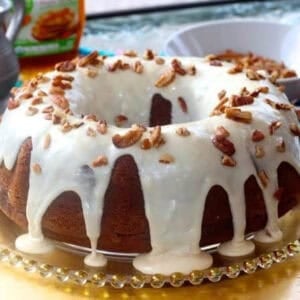 Pumpkin Bundt Cake topped with Maple Glaze and chopped pecans on a decorative cake plate next to a bowl of pecans and a bottle of maple syrup