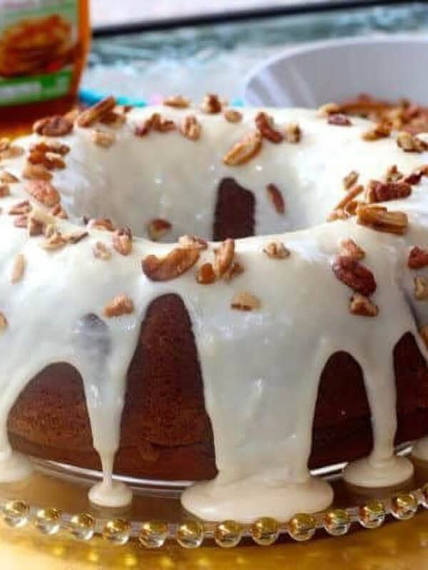 Pumpkin Bundt Cake topped with Maple Glaze and chopped pecans on a decorative cake plate next to a bowl of pecans and a bottle of maple syrup
