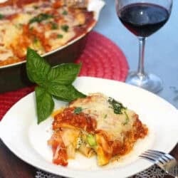 A slice of Pumpkin Lasagna on a plate garnished with four large basil leaves next to the remainder of the lasagna in a baking dish and a glass of red wine