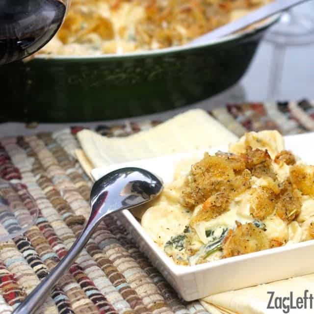 A scoop of Macaroni and Cheese with Kale and Gruyere topped with breadcrumbs on a square plate with a spoon resting on the side of the plate next to the remainder of the casserole in a baking dish