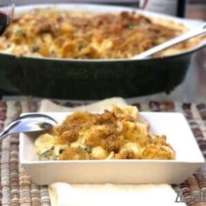A scoop of Mac and Cheese with Gruyere and kale topped with breadcrumbs on a square plate with a spoon resting on the side of the plate and the remainder of the casserole in a baking dish in the background