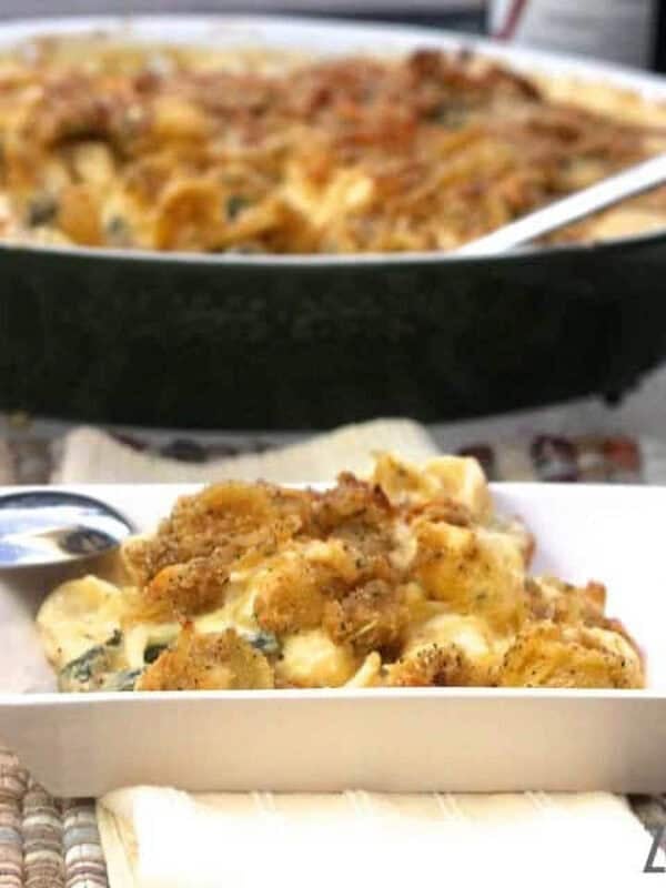 A scoop of Mac and Cheese with Gruyere and kale topped with breadcrumbs on a square plate with a spoon resting on the side of the plate and the remainder of the casserole in a baking dish in the background