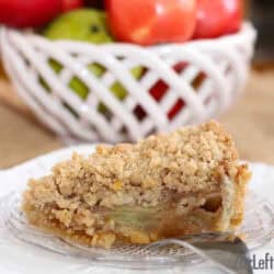 A slice of Caramel Apple Pie with Sugar Cookie Crust and streusel topping on a small plate with a large bowl of red and green apples in the background