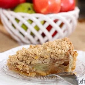 A slice of Caramel Apple Pie with Sugar Cookie Crust and streusel topping on a small plate with a large bowl of red and green apples in the background