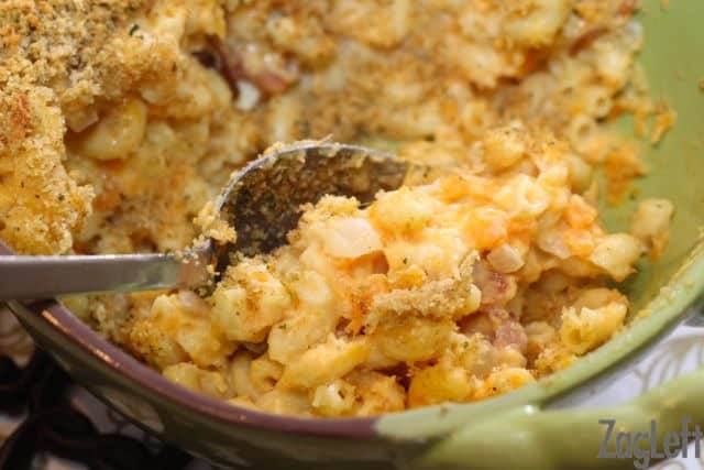 A closeup of a large spoon scooping Sweet Potato Mac and Cheese from a baking dish