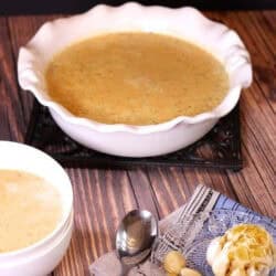 A large serving bowl of Roasted Garlic and Brie Soup next to a small bowl of soup and a small plate of roasted garlic and a spoon