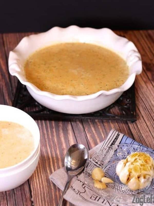 A large serving bowl of Roasted Garlic and Brie Soup next to a small bowl of soup and a small plate of roasted garlic and a spoon