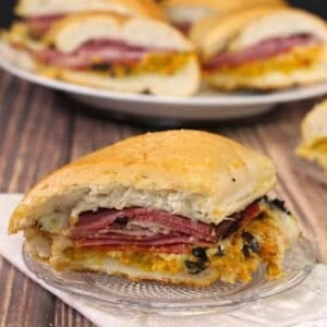 Two Muffuletta wedges on small plates with four more muffuletta wedges on a large plate in the background