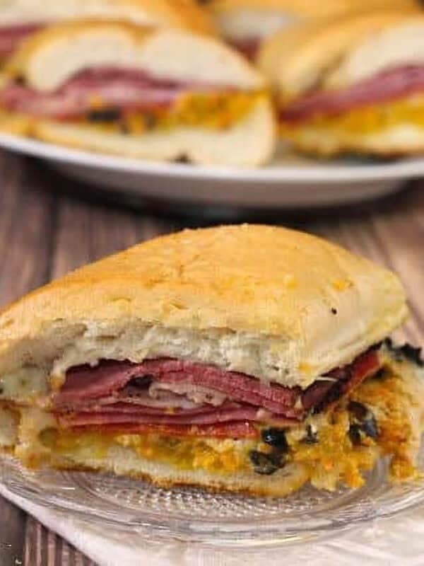 Two Muffuletta wedges on small plates with four more muffuletta wedges on a large plate in the background