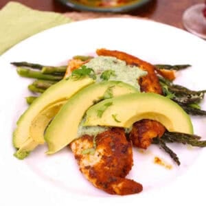 Blackened Tilapia topped with Cilantro Lime Sauce and three avocado slices on a bed of asparagus on a large plate next to half of an avocado and two limes on a small plate