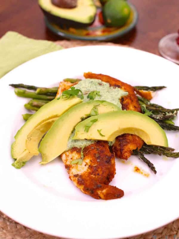 Blackened Tilapia topped with Cilantro Lime Sauce and three avocado slices on a bed of asparagus on a large plate next to half of an avocado and two limes on a small plate