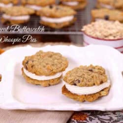 Two Oatmeal Butterscotch Whoopie Pies on a glass serving dish with whoopie pies on a cooling rack in the background next to a measuring cup of oats