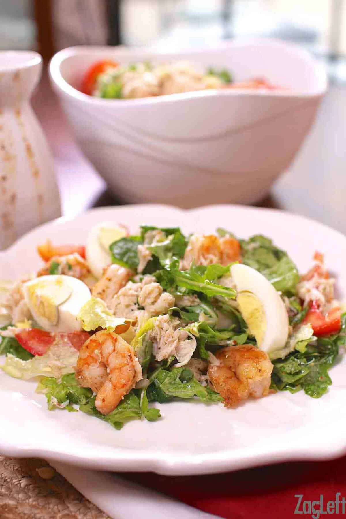 Shrimp and Crabmeat Salad topped with hard-boiled egg pieces on a large plate next to a serving bowl with the remainder of the salad