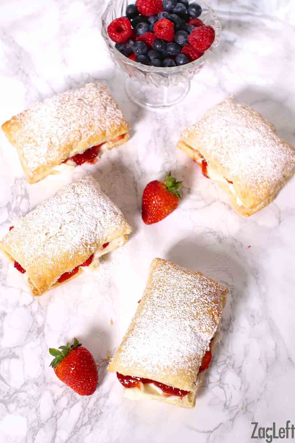 Overhead view of four strawberry cheesecake puffs dusted with powdered sugar next to a small bowl of raspberries and blueberries