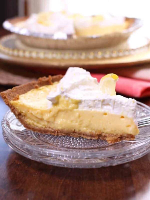Lemon Mango Icebox Pie topped with whipped cream and lemon slices on a large plate next to two whole lemons, a stack of forks, and a stack of small plates
