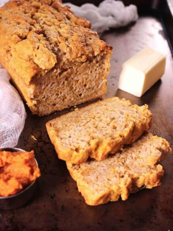 A loaf of Pumpkin Sage Beer Bread with two slices next to a small bowl of pumpkin puree and half a stick of butter all on a metal tray