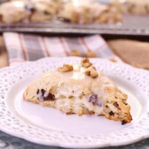 A Fig and Walnut Scone topped with a sweet Maple Glaze and chopped walnuts on a small plate, with scones on a cooling rack in the background