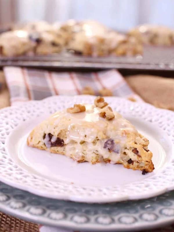 A Fig and Walnut Scone topped with a sweet Maple Glaze and chopped walnuts on a small plate, with scones on a cooling rack in the background