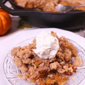 A slice of Crustless Pumpkin Pie with streusel topping and whipped cream on a small plate next to a cast iron skillet with the remainder of the pie