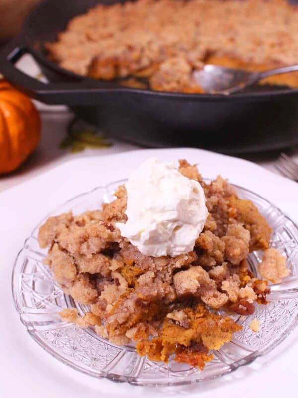 A slice of Crustless Pumpkin Pie with streusel topping and whipped cream on a small plate next to a cast iron skillet with the remainder of the pie