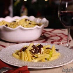 Mediterranean Pasta on a plate next to a large bowl of the remaining pasta and a glass of red wine