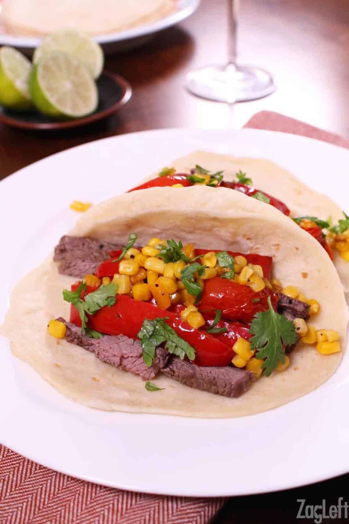 Two tacos with strips of steak, strips of red bell pepper, corn, and cherry tomatoes in a flour tortilla topped with cilantro on a plate with a small plate of lime halves in the background