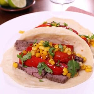 Two tacos with strips of steak, strips of red bell pepper, corn, and cherry tomatoes in a flour tortilla topped with cilantro on a plate with a small plate of lime halves in the background