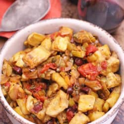 A bowl of Caponata next to a glass of red wine and a spoon all on a metal tray