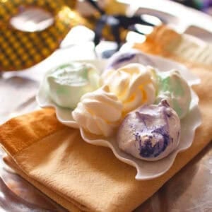 Six green, yellow, and purple Mardi Gras Meringues plated on a gold napkin on a metal tray with a gold Mardi Gras mask