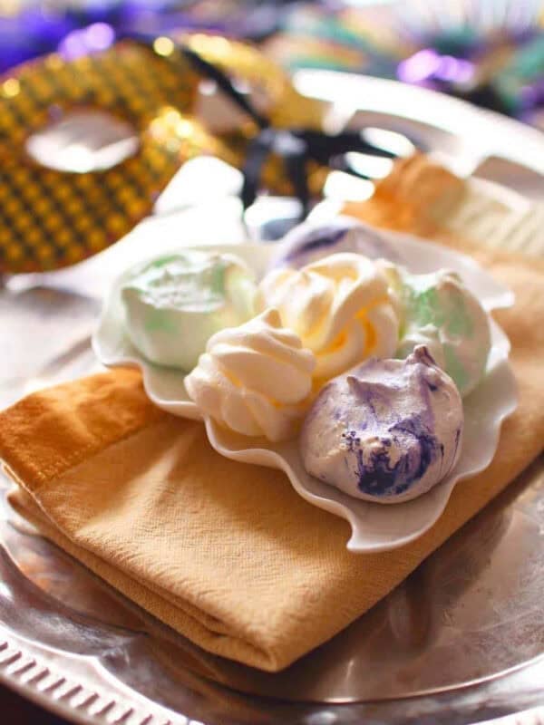 Six green, yellow, and purple Mardi Gras Meringues plated on a gold napkin on a metal tray with a gold Mardi Gras mask