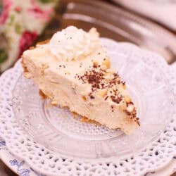 A slice of Peanut Butter Marshmallow Cream Pie topped with whipped cream and chocolate shavings plated on a metal tray with a floral cloth napkin and the remainder of the pie on a cake stand in the background