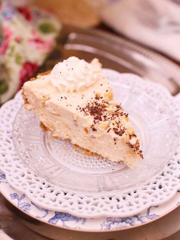 A slice of Peanut Butter Marshmallow Cream Pie topped with whipped cream and chocolate shavings plated on a metal tray with a floral cloth napkin and the remainder of the pie on a cake stand in the background