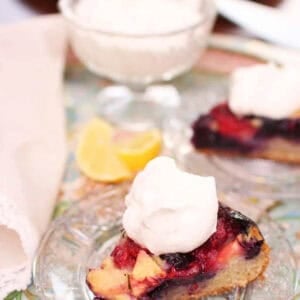 A slice of Apple Berry Coffee Cake topped with whipped cream on a small plate next to a dessert glass of whipped cream all on a metal tray