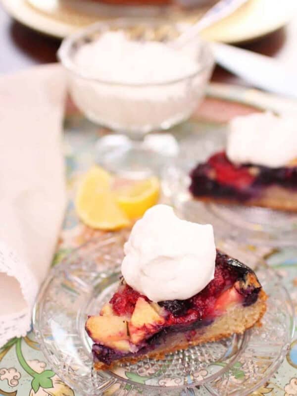 A slice of Apple Berry Coffee Cake topped with whipped cream on a small plate next to a dessert glass of whipped cream all on a metal tray