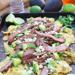 Thick tortilla chips lightly cooked in salsa topped with thinly sliced flank steak, avocados, Cotija cheese and cilantro on a baking sheet