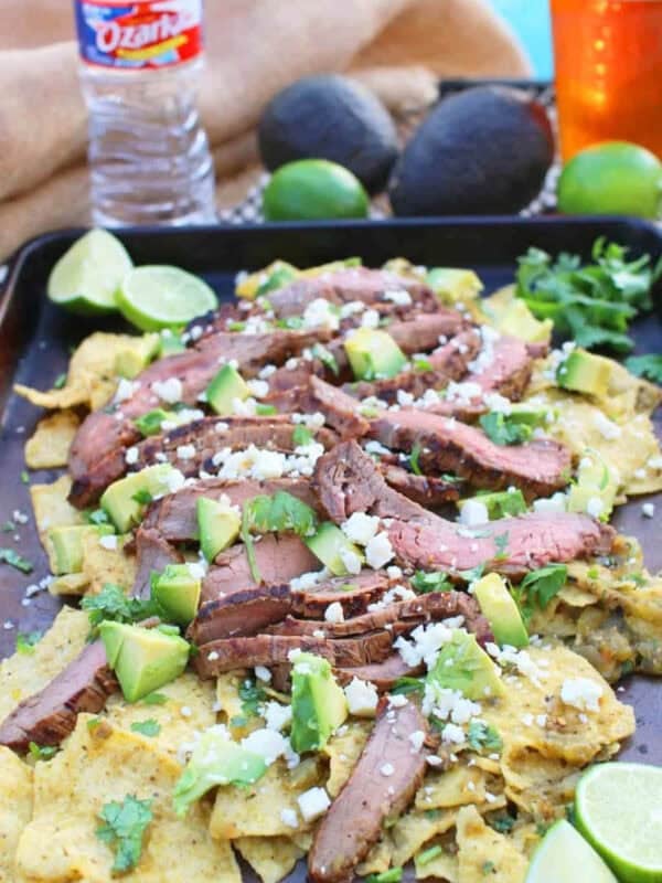 Thick tortilla chips lightly cooked in salsa topped with thinly sliced flank steak, avocados, Cotija cheese and cilantro on a baking sheet