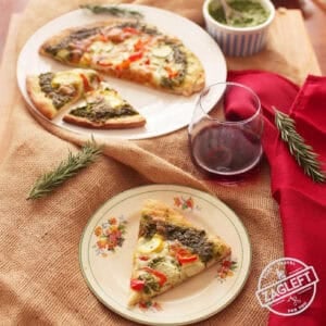 Overhead view of a small plated slice of pesto pizza and the rest of the pizza on a larger plate next to a glass of red wine and a small bowl of pesto all on a burlap fabric cloth on a wooden table with two sprigs of rosemary and a red napkin