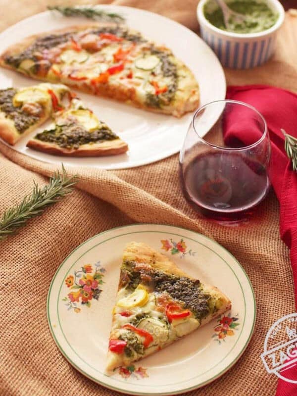 Overhead view of a small plated slice of pesto pizza and the rest of the pizza on a larger plate next to a glass of red wine and a small bowl of pesto all on a burlap fabric cloth on a wooden table with two sprigs of rosemary and a red napkin