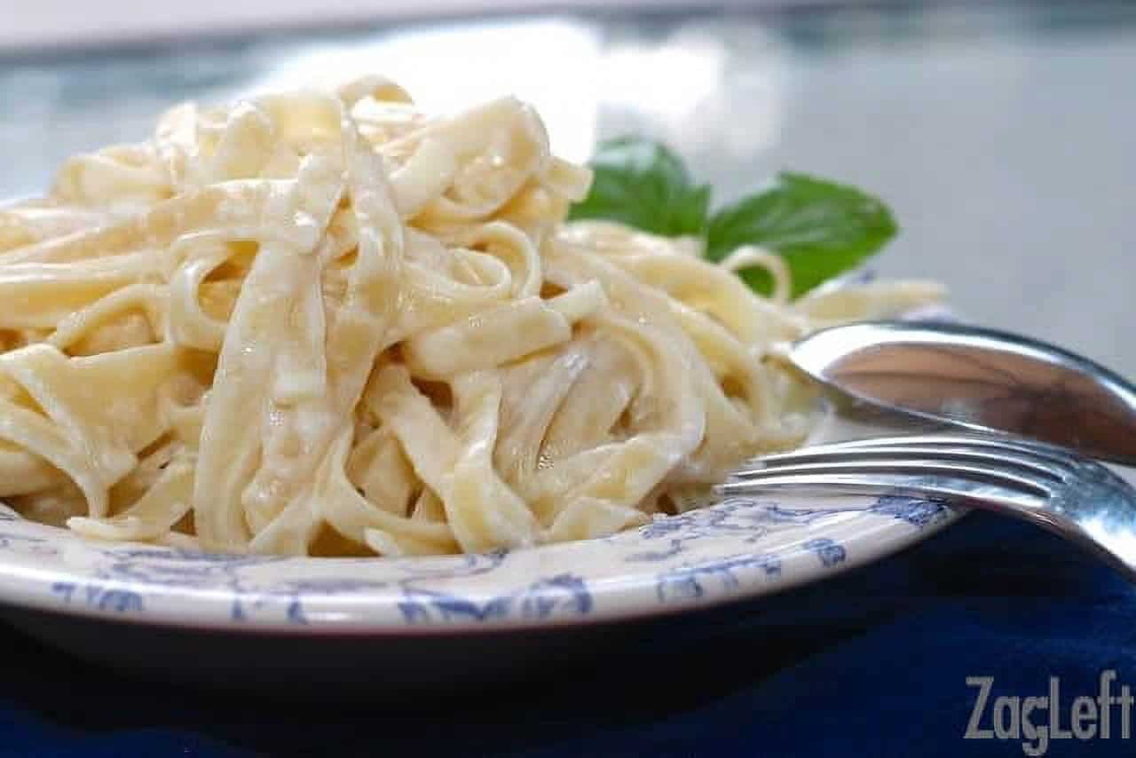 A plate of pasta with Alfredo sauce with a fork and spoon resting on the side of the plate.