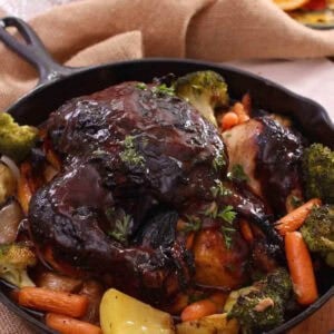 Baked chicken covered with bbq sauce with baby carrots, broccoli florets, and potatoes in a cast iron skillet with a small bowl of white rice and a small plate of orange slices in the background