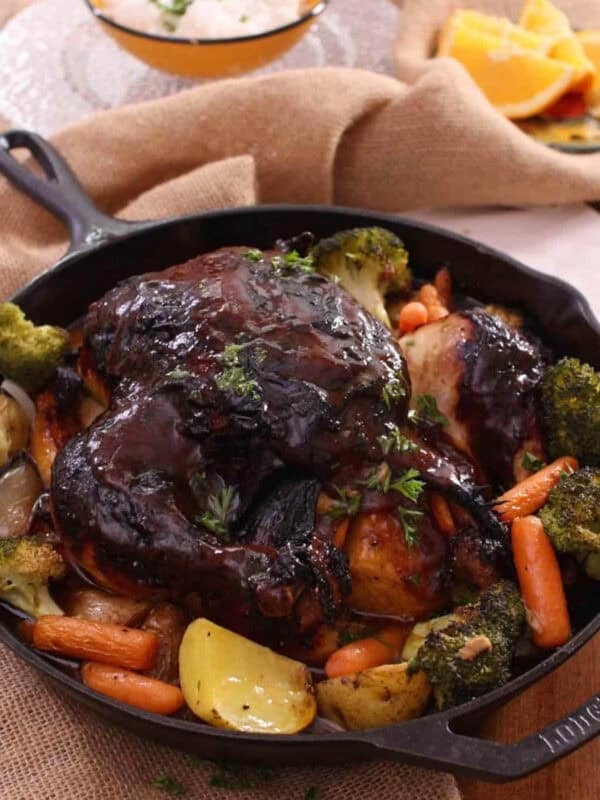 Baked chicken covered with bbq sauce with baby carrots, broccoli florets, and potatoes in a cast iron skillet with a small bowl of white rice and a small plate of orange slices in the background